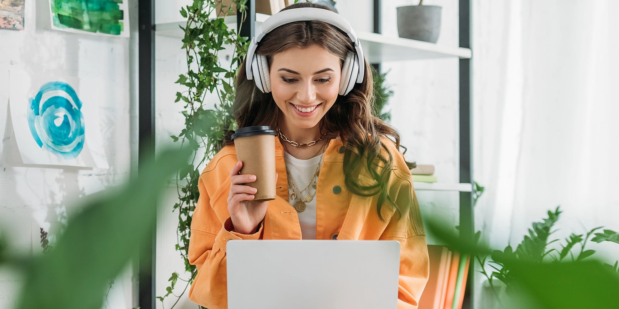 Lächelnde Frau an Laptop im Office mit Pflanzen und Kaffee in der Hand